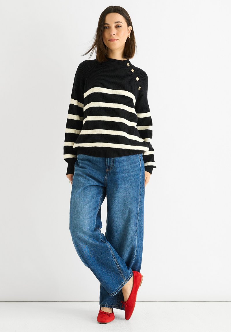Black and white striped sweater with a ribbed texture and button details, paired with loose blue jeans and red flat shoes. White background.