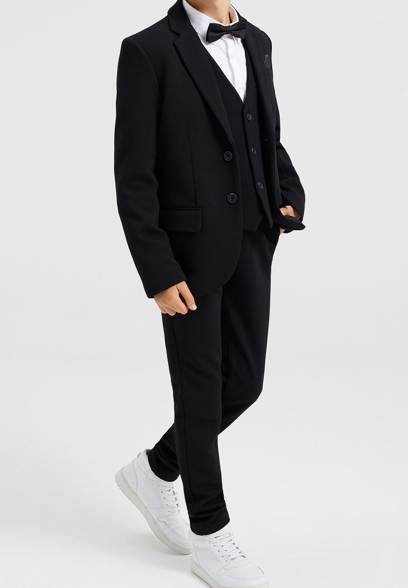 Costume trois pièces noir avec un blazer texturé, un gilet assorti et un pantalon sur mesure, associé à une chemise blanche et un nœud papillon noir. Baskets blanches.