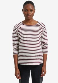 Frau trägt ein langärmliges weißes Hemd mit bordeauxfarbenen horizontalen Streifen und Knopfdetails am Hals, kombiniert mit schwarzen Hosen.