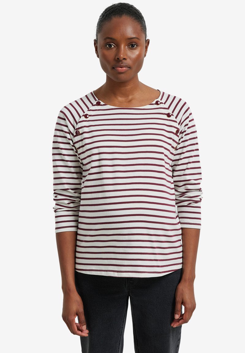 Frau trägt ein langärmliges weißes Hemd mit bordeauxfarbenen horizontalen Streifen und Knopfdetails am Hals, kombiniert mit schwarzen Hosen.