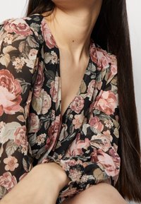 Blusa floral con fondo negro y rosas en tonos rosa y crema, fabricada en tejido semitransparente, diseño de escote en V y puños elásticos.