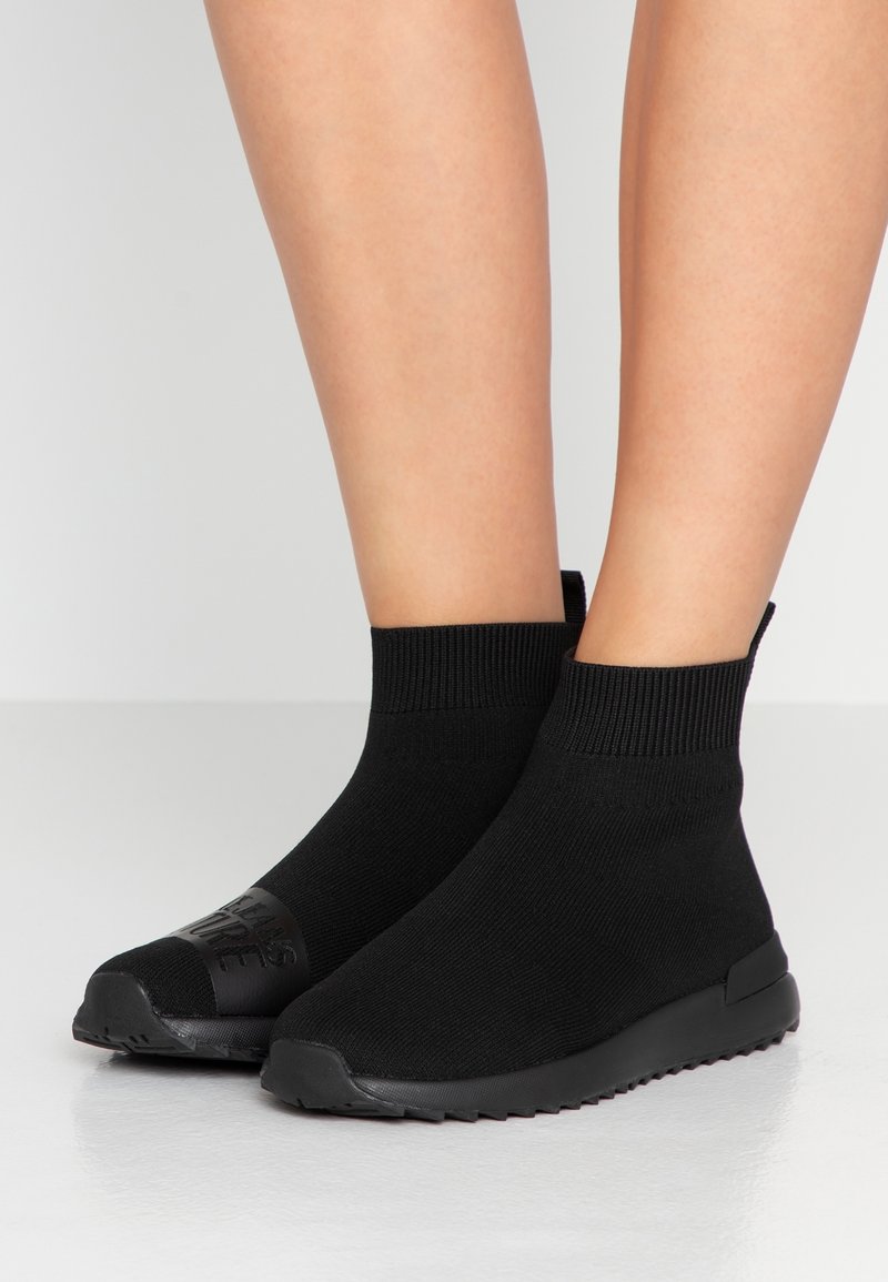 Bottines en maille noire à texture lisse, avec des poignets élastiques et des semelles en caoutchouc. Présentent un logo embossé sur le bout de la chaussure.
