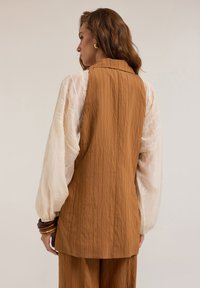 Chaleco marrón texturizado con cuello en la parte posterior, superpuesto a una blusa crema con detalles bordados y mangas abullonadas.