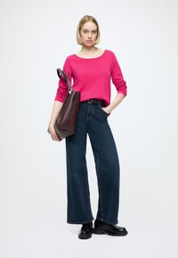 Dolga roza majica z dolgimi rokavi v kombinaciji s širokimi hlačami iz temnega jeansa. Dodatki vključujejo teksturirano bordo torbo za na ramo. Črna robustna obutev dopolnjuje videz.