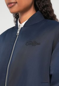 Temno modra bomber jakna z rebrastim ovratnikom, srebrnim zadrgo in vezenim logotipom na prsih. Gladek, lahek material.