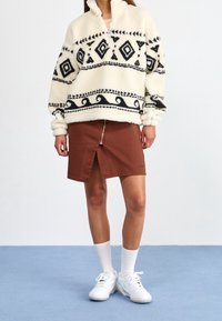 Krémový fleecový pulovr s černým diamantovým vzorem, design s polovičním zipem; doplněný hnědou sukní s předním zipem a rozparkem. Bílé ponožky a boty.