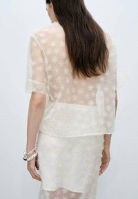 Blusa bianca trasparente con ricami di margherite gialle, maniche corte e vestibilità ampia, abbinata a una gonna trasparente coordinata. Accessorio trasparente al polso.