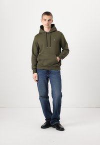 Oliven grønn hettegenser med kengurulomme, snor i hetta, og logodetalj. Kombinert med blå denimjeans og svarte sko.