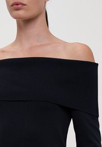 Haut noir à épaules dénudées avec une texture côtelée, coupe ajustée et un large décolleté, mettant en valeur la peau des épaules et de la clavicule.