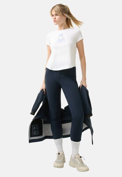 White short-sleeve t-shirt with graphic design, paired with navy athletic pants and light-colored shoes. Jacket held in one hand; simple design.