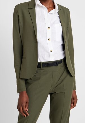 Person, die einen olivgrünen Blazer und passende Hose, ein weißes Hemd und einen schwarzen Gürtel trägt, steht vor einem schlichten hellen Hintergrund.