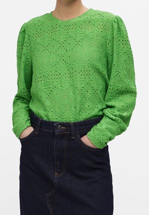 Personne portant un chemisier en dentelle vert vif à manches longues avec des épaules bouffantes et une jupe en jean bleu marine taille haute, mains dans les poches.