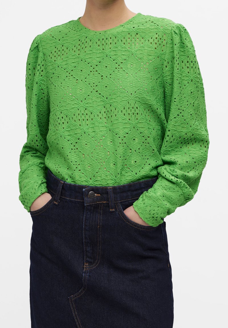 Personne portant un chemisier en dentelle vert vif à manches longues avec des épaules bouffantes et une jupe en jean bleu marine taille haute, mains dans les poches.