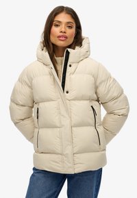 Veste d'hiver beige avec capuche, dotée d'une fermeture éclair à l'avant, d'accents noirs et de deux poches latérales. Texture matelassée avec un motif en losanges.