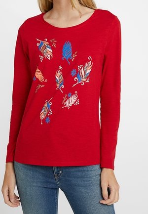 Chemise rouge à manches longues avec des motifs colorés de plumes et de fleurs sur le devant, portée avec un jean bleu.