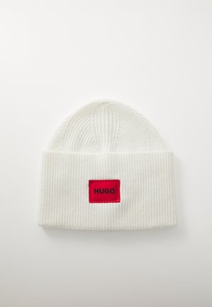 Witte geribbelde gebreide beanie met een omgevouwen rand en een rechthoekige rode patch met "HUGO" in zwarte tekst. Zachte textuur, ronde top.