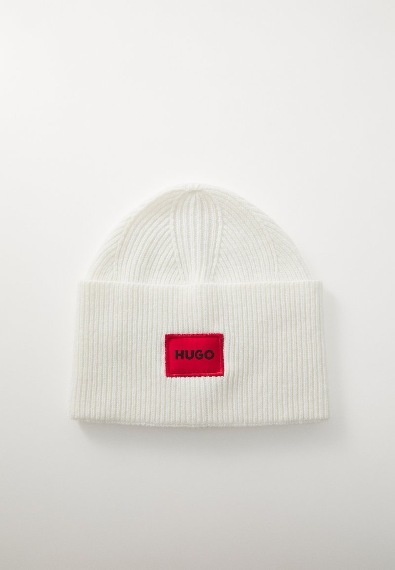 White ribbed knit beanie featuring a folded cuff and a rectangular red patch with "HUGO" in black text. Soft texture, rounded top.