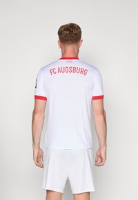 Hvit fotballtrøye med røde detaljer og "FC Augsburg" i fet rød skrift på ryggen. Korte ermer, lett stoff.