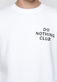 Bílá bavlněná mikina s kulatým výstřihem, s černým vyšívaným textem "DO NOTHING CLUB" na levé části hrudi. Hladká textura.