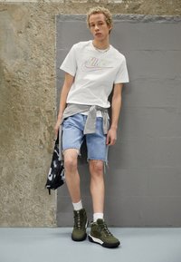 T-shirt Nike blanche avec un logo coloré, pull gris noué autour de la taille, shorts en denim décolorés et baskets montantes vertes. Sac noir et blanc.