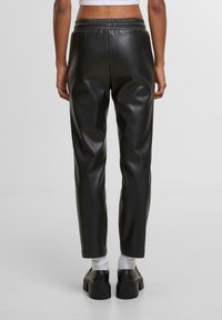 Černé kalhoty z umělé kůže, tapered střih, s elastickým pasem, hladkou texturou a do kotníků, doplněné o robustní černé boty a bílé ponožky.