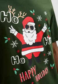 Camiseta de algodón verde con un Papá Noel de dibujos animados usando gafas de sol, traje rojo y barba blanca, rodeado de copos de nieve y acebo. El texto dice "Ho Ho Felices Fiestas".