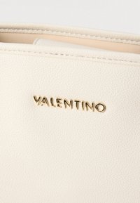 Kremfarget skinnveske med teksturert overflate, med en metallisk gull "VALENTINO" logo. Designet er minimalistisk med synlige søm-detaljer.