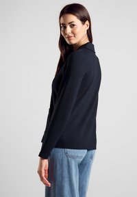 Schwarzer Blazer mit langen Ärmeln, ausgefallenem Revers, glattem Stoff und zwei Fronttaschen. Kombiniert mit hellblauen Jeans.