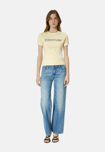 Young woman wearing a yellow short-sleeve shirt with "KOOPLES" text, wide-leg blue jeans, and black strappy sandals, standing facing forward.