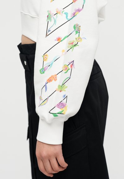 Λευκό φούτερ με πολύχρωμη κεντητή λουλουδάτη διακόσμηση κατά μήκος του μανικιού, με μαύρες λεπτομέρειες που δημιουργούν γεωμετρικά σχήματα. Διάφανα μαύρα παντελόνια με τσέπες.