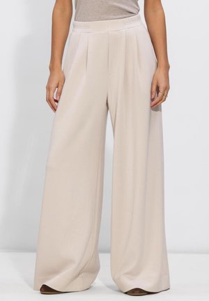 Beige High-Waist-Hose mit weitem Bein und vorderen Falten, getragen mit einem in die Hose gesteckten hellbeigen Oberteil und braunen Schuhen.