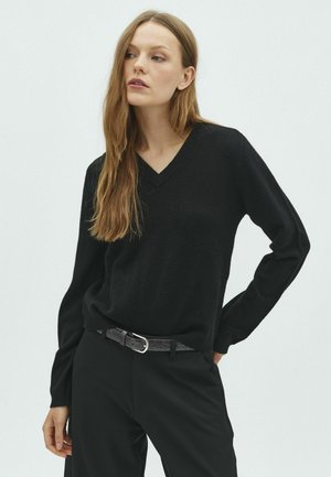 Femme aux longs cheveux châtain clair portant un pull noir à col en V, un pantalon noir et une ceinture noire, debout avec une main sur la hanche.