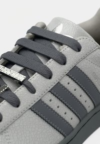 Γκρι δερμάτινο αθλητικό παπούτσι με υφή, σκουρόγκρι κορδόνια και τρεις σκουρόγκρινες ρίγες. Διαθέτει μεταλλικό λογότυπο στη γλώσσα του παπουτσιού.