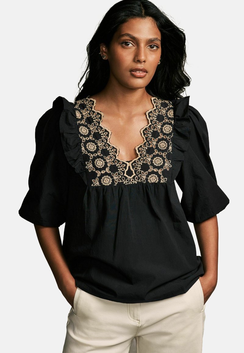 Blusa nera con maniche corte a sbuffo, scollo arricciato e complessa ricamatura floreale beige lungo il colletto. Tessuto di cotone liscio.