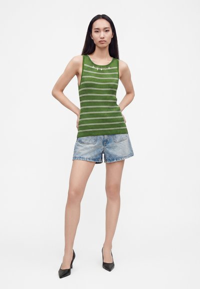 Femme aux longs cheveux noirs portant un haut rayé vert sans manches, un short en jean et des chaussures noires à talons hauts, debout les mains sur les hanches.