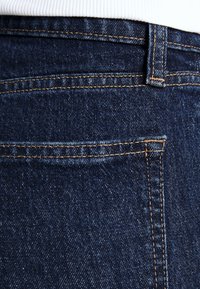 Dark blue denim jeans showing back pocket with orange stitching and a belt loop, white fabric visible at the top.