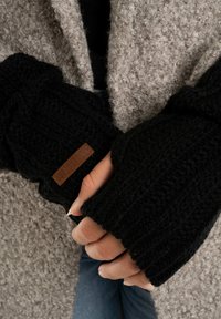 Zwarte gebreide wanten zonder vingers met ribbeltextuur en een leren label, gedragen over een lichtgrijze, fuzzy buitenstof.