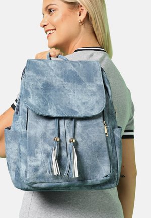 Denim-agtig blå rygsæk med klap, to frynser og sidelommer. Har en struktureret overflade og skinnende metal-accenter.
