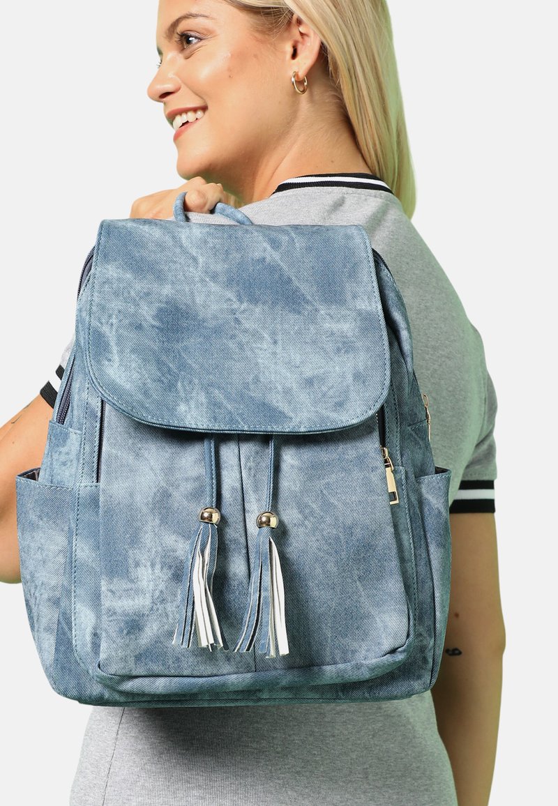 Modrý batoh ve stylu džínoviny s klopou, dvěma třásněmi a bočními kapsami. Má texturovaný povrch a lesklé kovové akcenty.