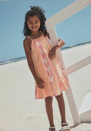 Young girl in a patterned pink and orange sundress leaning on a white wooden structure on a sandy beach under clear blue sky.