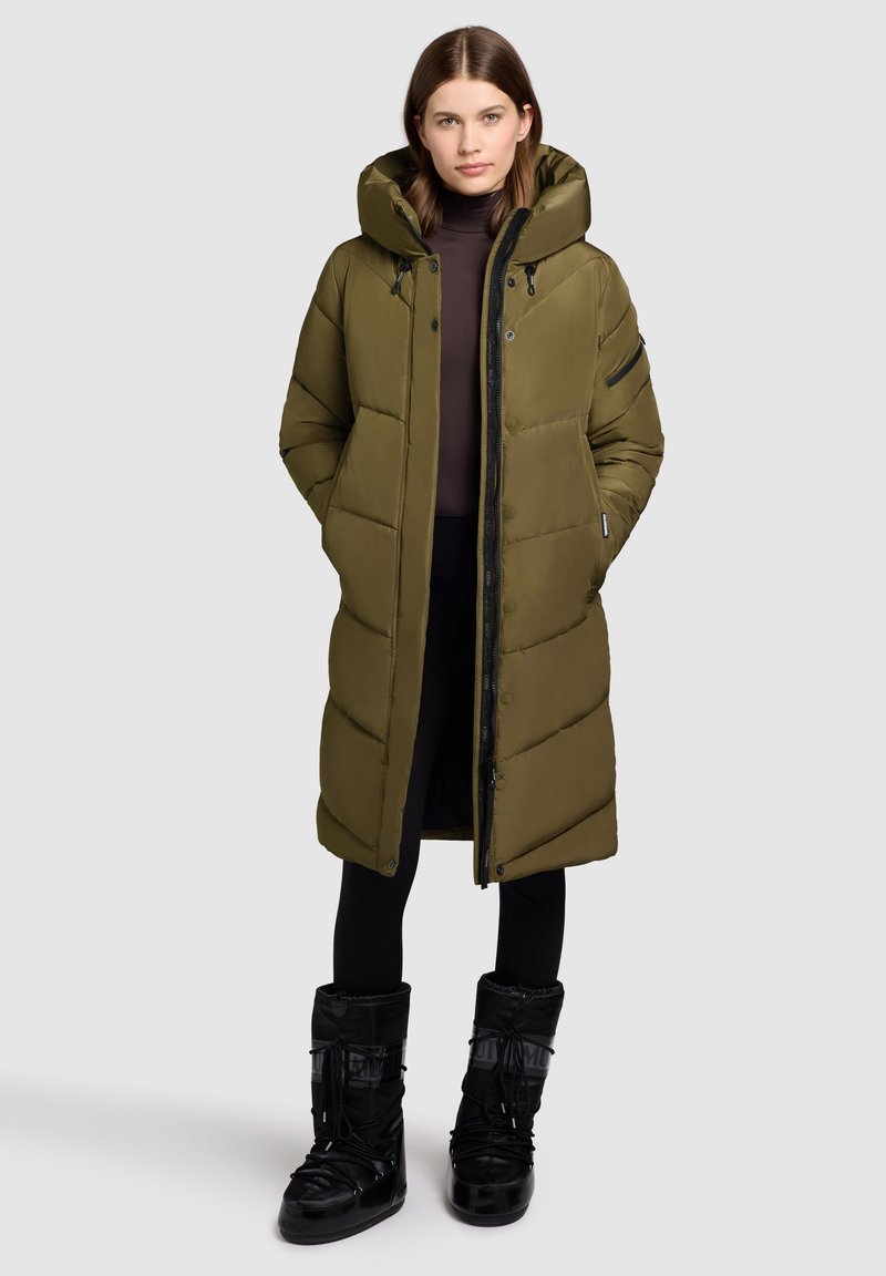 Cappotto lungo imbottito verde oliva con cappuccio, zip frontale, tasche laterali e tessuto testurizzato; abbinato a stivali neri con lacci e dettagli.