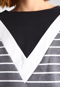 Gestreifter grauer und weißer Stoff mit einem schwarzen V-Ausschnitt-Kragen. Das Design zeigt kräftige dreieckige Muster, wo die Farben aufeinandertreffen.