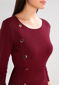 Burgundy pitkähihaine paita ribbittyöstä, jossa on pyöreä kaula-aukko ja kultaiset napit vasemmalla puolella.