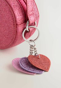 Kulatá taška s růžovým třpytem na zip se stříbrnými kovovými doplňky, která obsahuje přívěsek se třemi srdíčkovými štítky v růžové, fialové a červené třpytivé barvě.