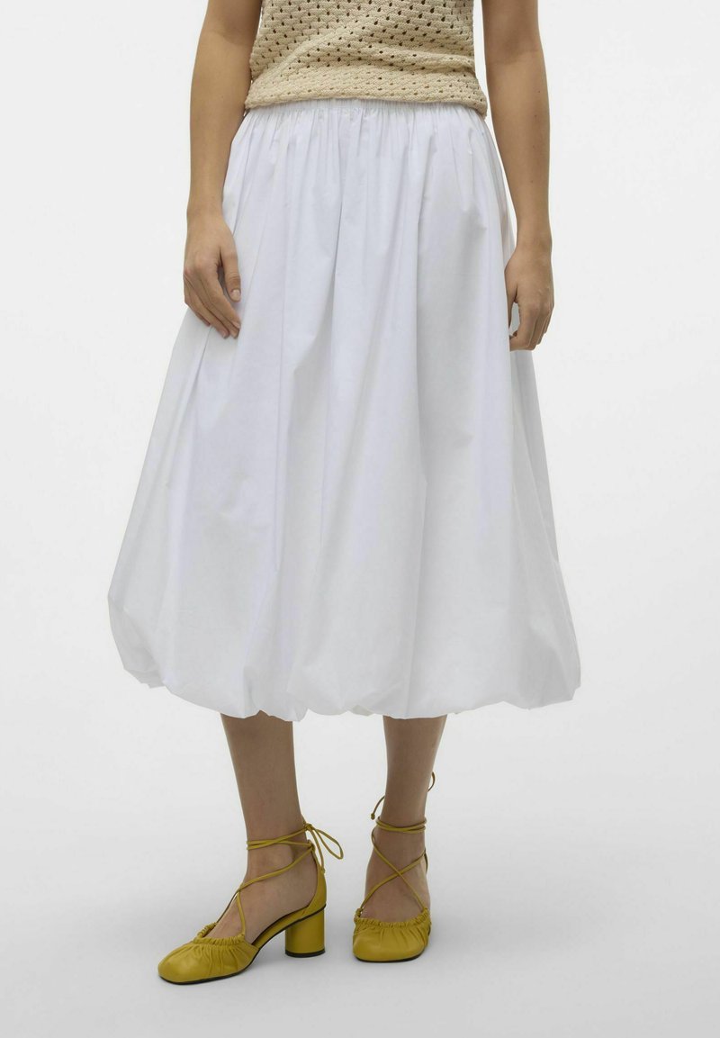 Vrouw in een witte blote midi-rok, beige haak-top en mosterdgele gestrikte blokhak-schoenen staande op een effen achtergrond.