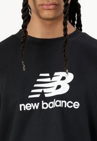 Črna majica s kratkimi rokavi z belim logotipom "New Balance", ki vsebuje stilizirano 'N' v gibanju. Narejena iz mehkega materiala s krojnim izrezom.