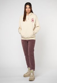 Frau steht mit den Händen in den Taschen, trägt einen cremefarbenen Hoodie mit Grafik, rosa Cordhosen und beigen klobigen Stiefeln vor einem einfarbigen Hintergrund.