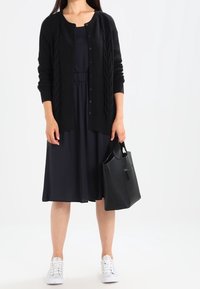 Svart stickad cardigan med kabelmönster, bärs över en marinblå klänning. Matchas med vita sneakers och en svart handväska. Slät tygtextur.