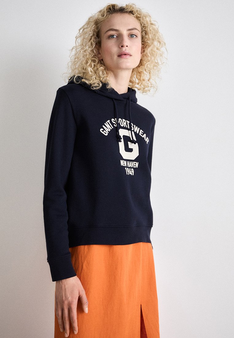 Navy Hoodie mit weißem Logo und Text, ausgestattet mit einer Kapuze mit Kordelzug und gerippten Bündchen. Kombiniert mit einem leuchtend orangefarbenen Rock mit Schlitz.