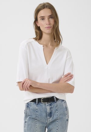 Mujer con cabello hasta los hombros, vistiendo una camiseta blanca de manga corta con cuello en V y jeans azul claro con cinturón negro, con los brazos cruzados.